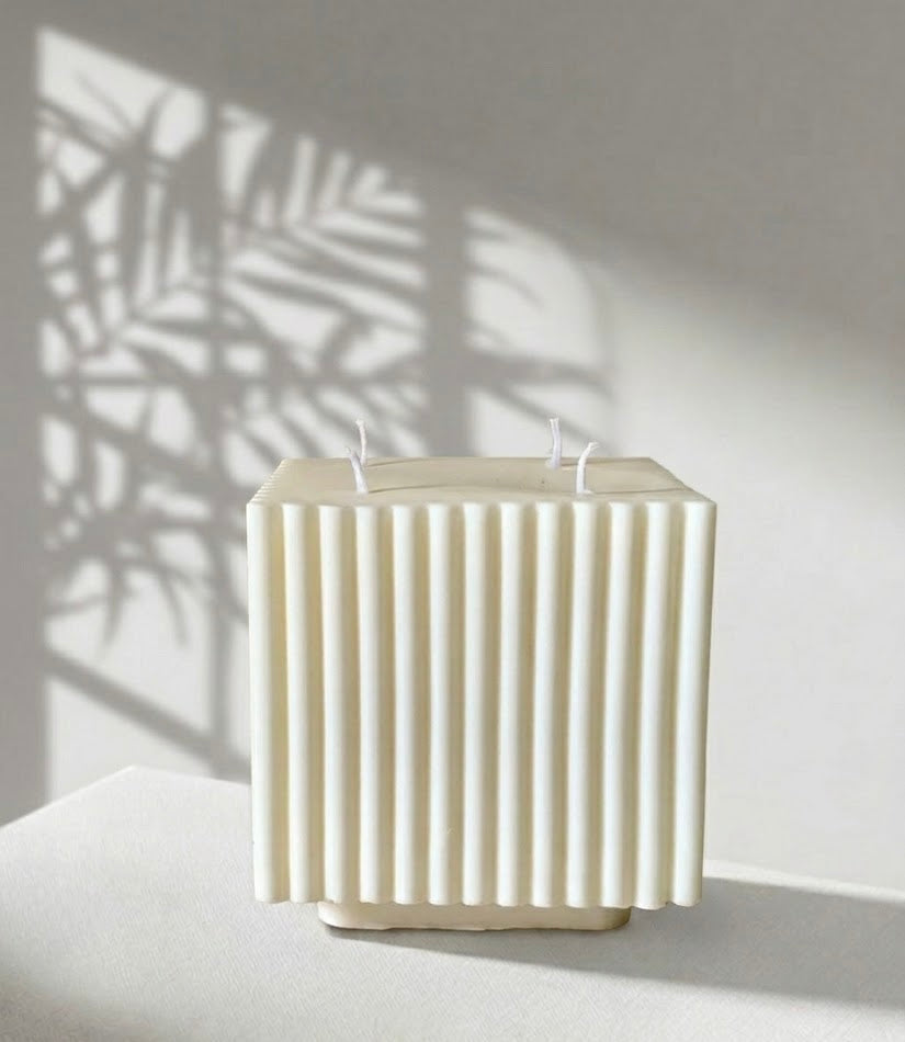 White textured candle in a rectangular holder on a light surface with a shadow of leaves on the wall.